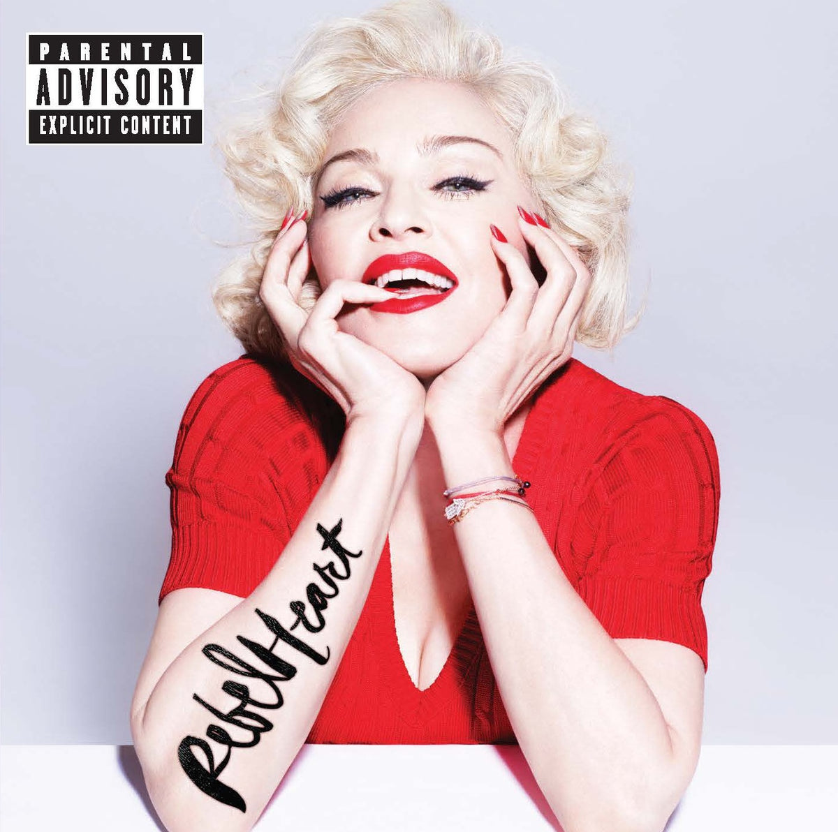 Madonna wearing a red top with 'Rebel Heart' tattoo on arm, against a plain background.